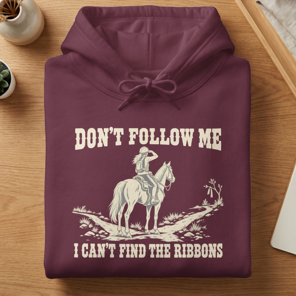 Maroon hoodie folded on a wooden table, featuring graphic text reading Don't follow me I can't find the ribbons and a horse rider illustration.