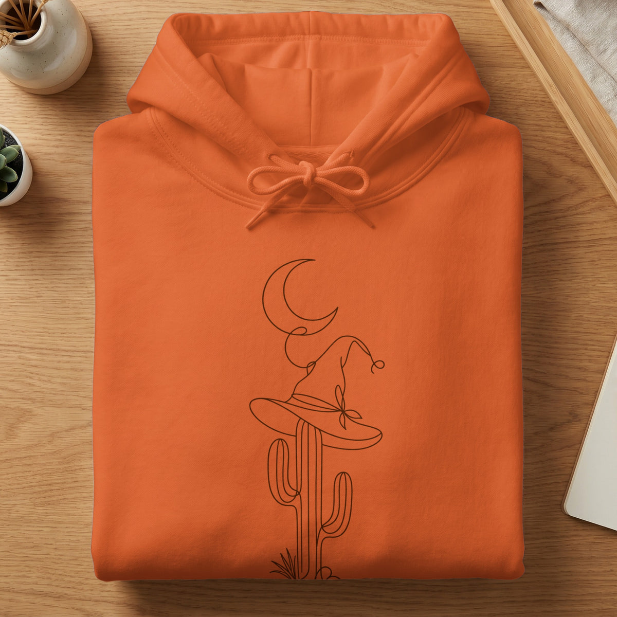 Orange hoodie with a line drawing of a cactus wearing a witch hat under a moon.