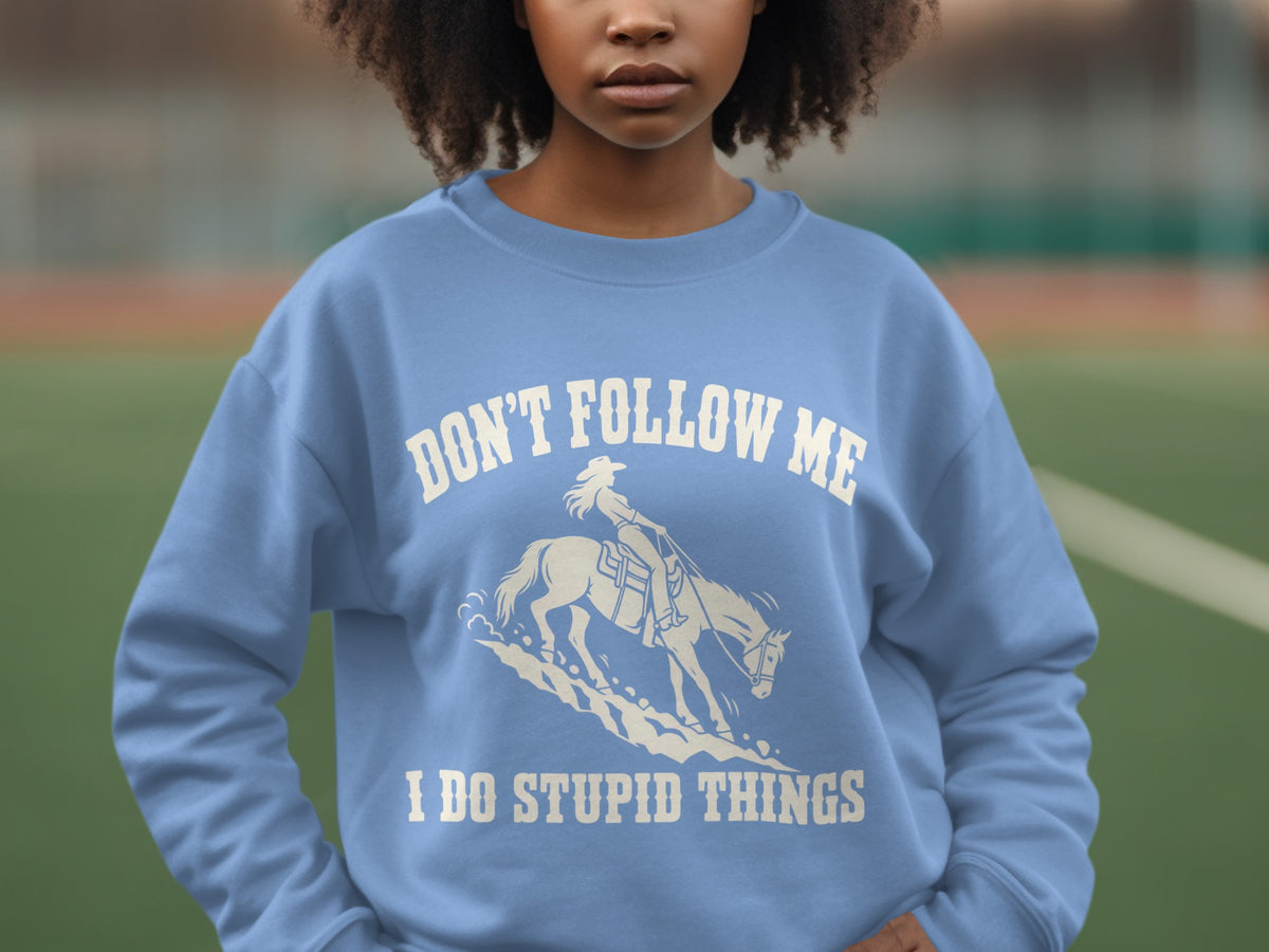 A person wearing a light blue sweatshirt featuring white text reading DON'T FOLLOW ME I DO STUPID THINGS and an illustration of a woman riding a horse.