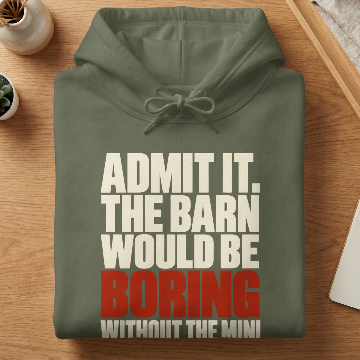 An olive green hoodie folded on a wooden surface with white and red text reading ADMIT IT. THE BARN WOULD BE BORING.