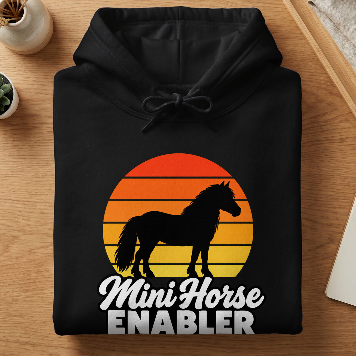 A folded black hooded sweatshirt featuring a retro sunset graphic and the text Mini Horse Enabler.