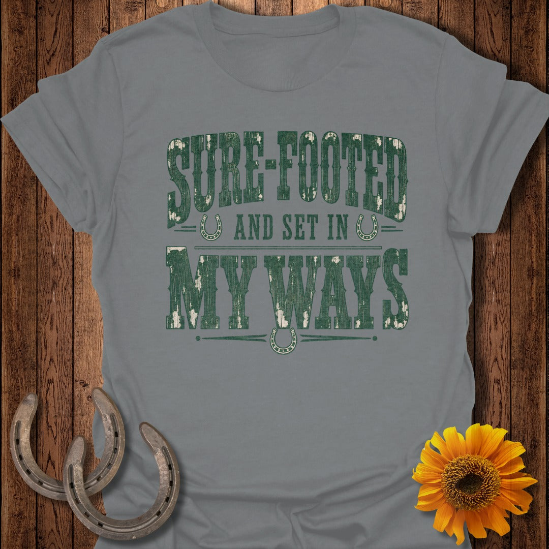 Gray t-shirt with distressed green text reading "SURE-FOOTED AND SET IN MY WAYS" with horseshoes.