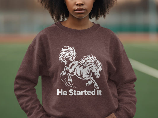A person wearing a brown sweatshirt with a white graphic of a running horse and the text He Started It.