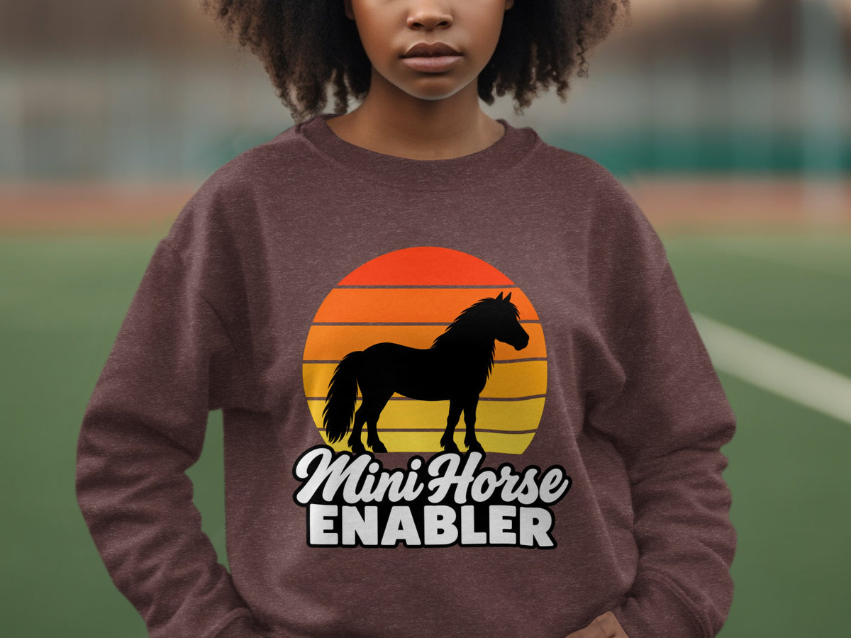 A person wearing a brown sweatshirt with a graphic of a horse and sunset and the text Mini Horse Enabler.