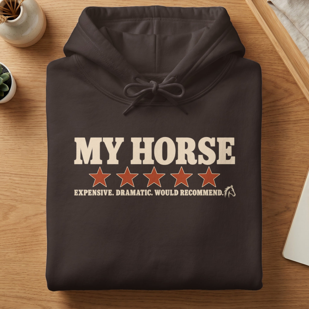A folded dark brown hoodie featuring cream and red text that reads MY HORSE, five stars, and EXPENSIVE. DRAMATIC. WOULD RECOMMEND.