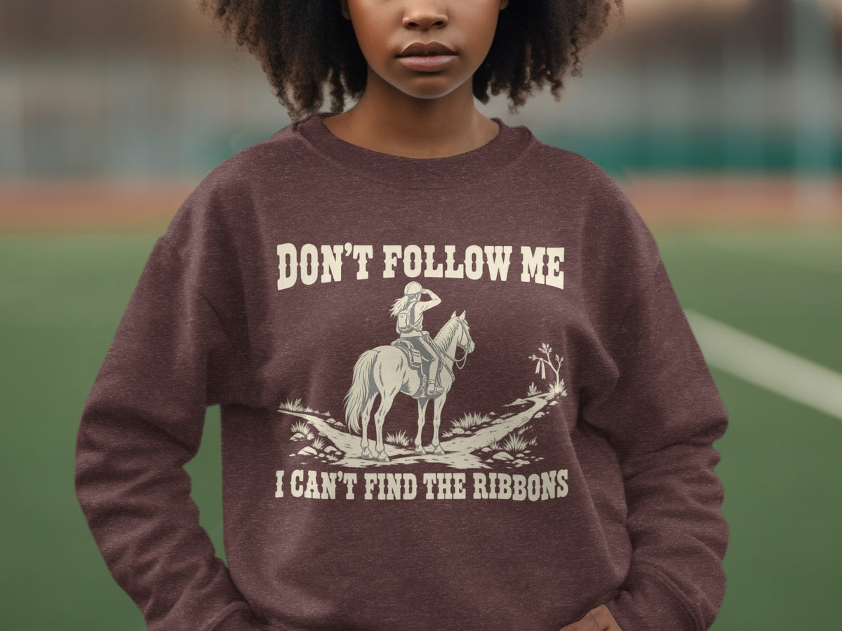 A person wearing a brown crewneck sweatshirt featuring a graphic of a horse rider and the text DON'T FOLLOW ME I CAN'T FIND THE RIBBONS.