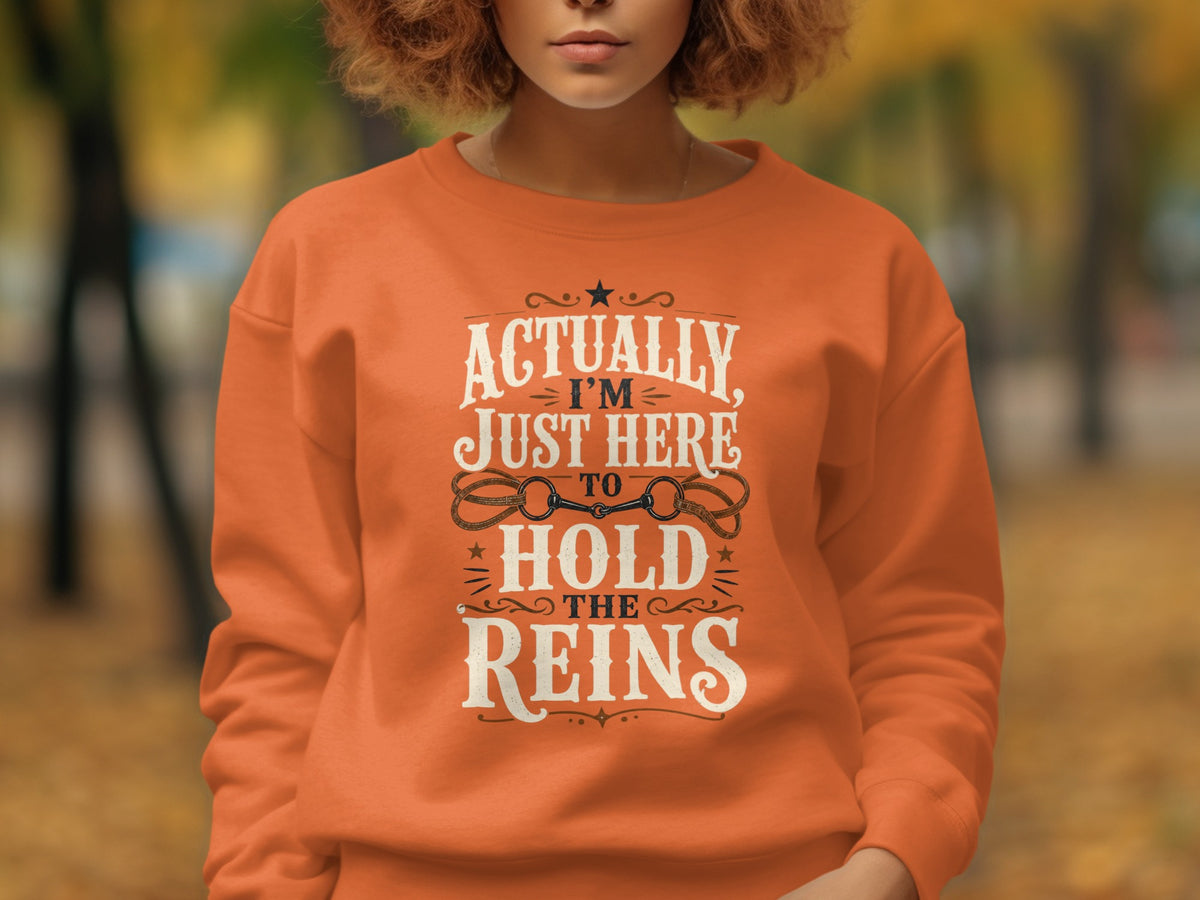 Orange sweatshirt with "Actually, I'm Just Here To Hold The Reins" text and horse tack design.