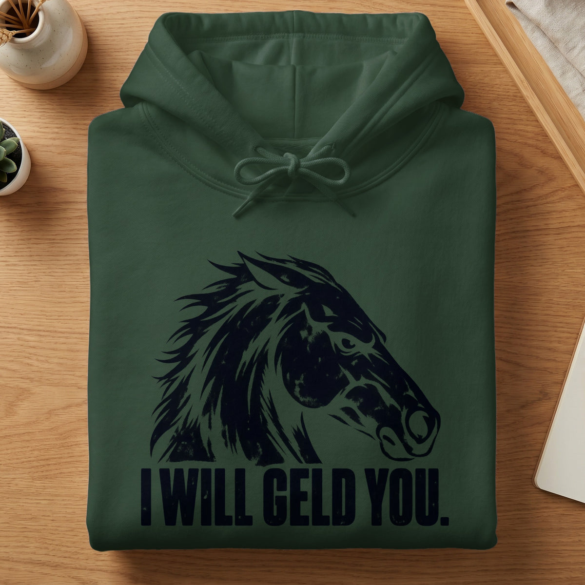 A folded dark green hoodie on a wooden table features a black horse graphic and the text I WILL GELD YOU.