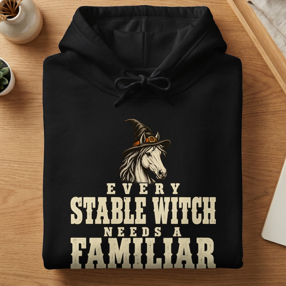 Black hoodie with white text reading "EVERY STABLE WITCH NEEDS A FAMILIAR" and a horse wearing a witch hat.