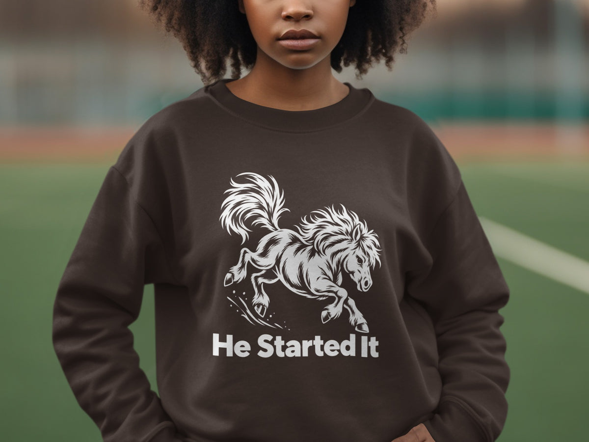 A person wearing a dark brown crewneck sweatshirt featuring a white graphic of a running horse and the text He Started It.