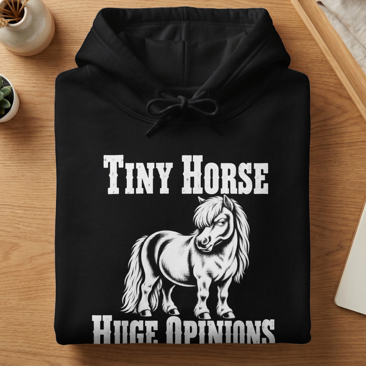 A folded black hooded sweatshirt with white text reading Tiny Horse Huge Opinions surrounding a pony illustration.