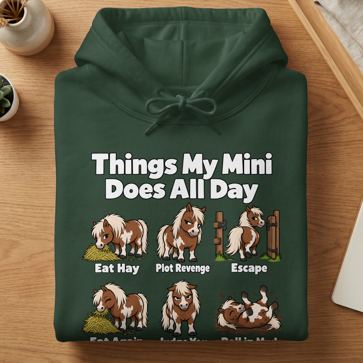 A folded dark green hooded sweatshirt features cartoon illustrations of a miniature horse and the text Things My Mini Does All Day.