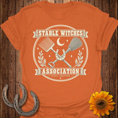 Orange t-shirt with "Stable Witches Association Local Chapter" graphic featuring a broom, pitchfork, and horseshoe.