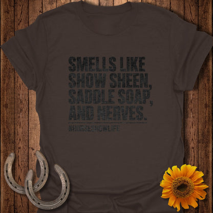 Brown crewneck t-shirt with black text reading "Smells Like Show Sheen, Saddle Soap, and Nerves." on a wooden background.