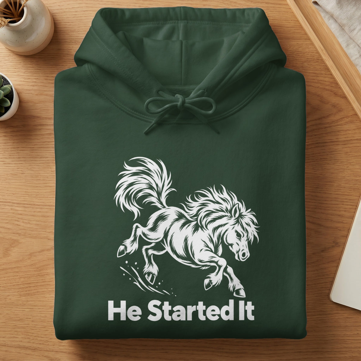 A folded dark green hooded sweatshirt featuring a white graphic of a galloping horse with the text He Started It underneath.