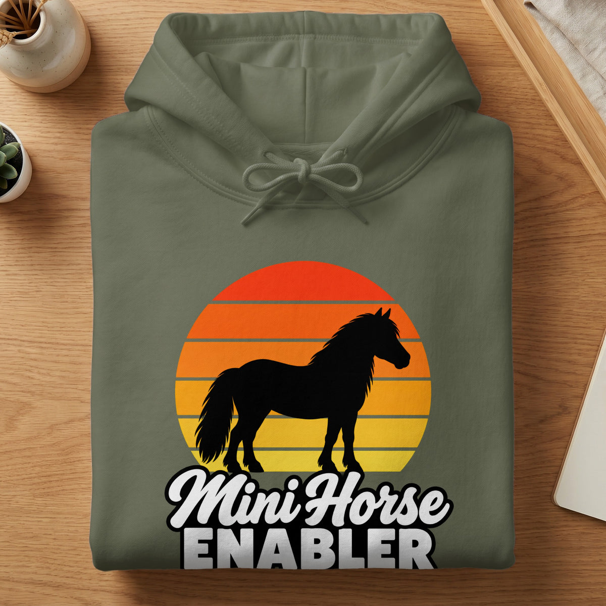 Folded olive green hoodie with a sunset silhouette graphic of a miniature horse and text reading Mini Horse Enabler.
