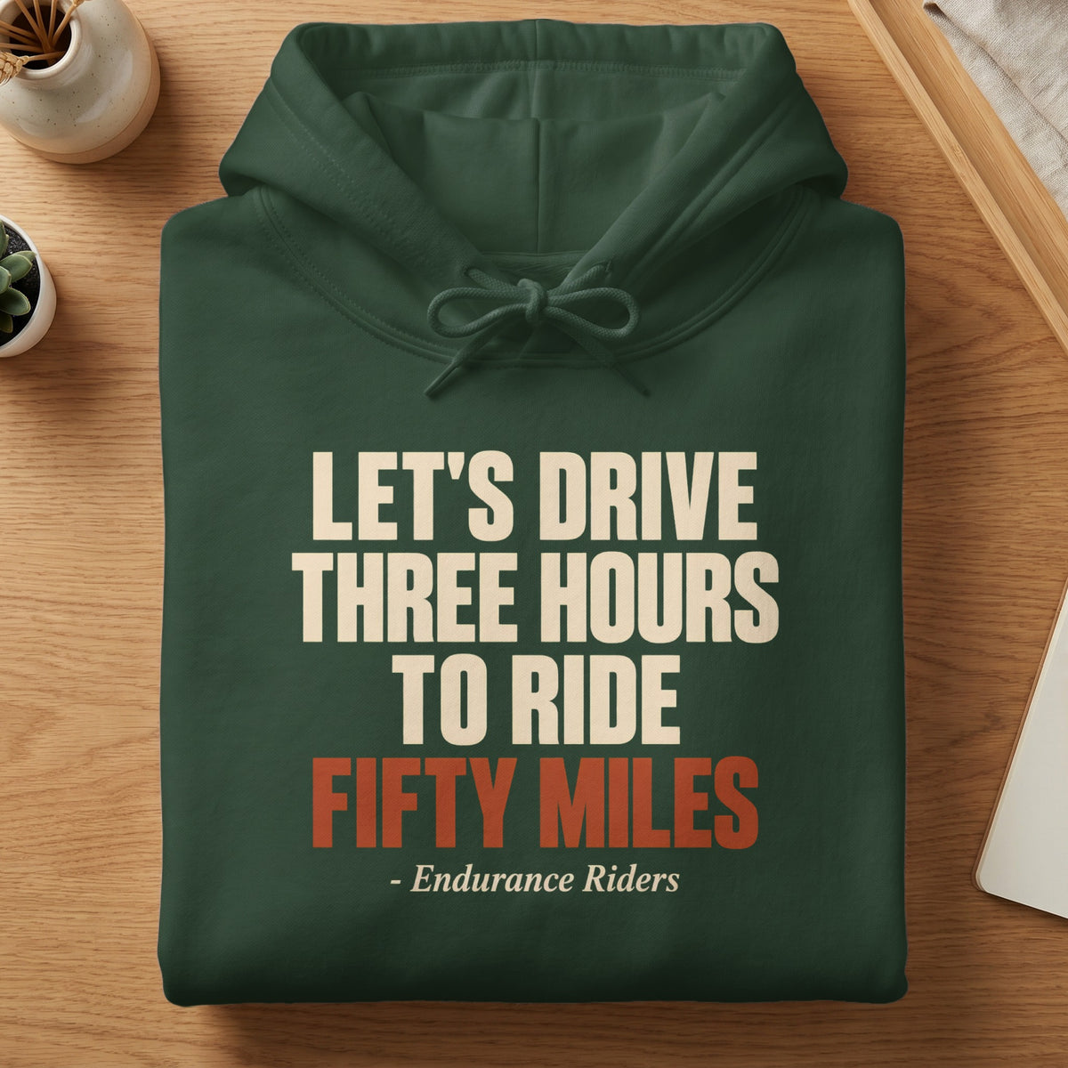 Folded dark green hooded sweatshirt with white and orange text reading "Let's Drive Three Hours to Ride Fifty Miles - Endurance Riders" on a wooden table.