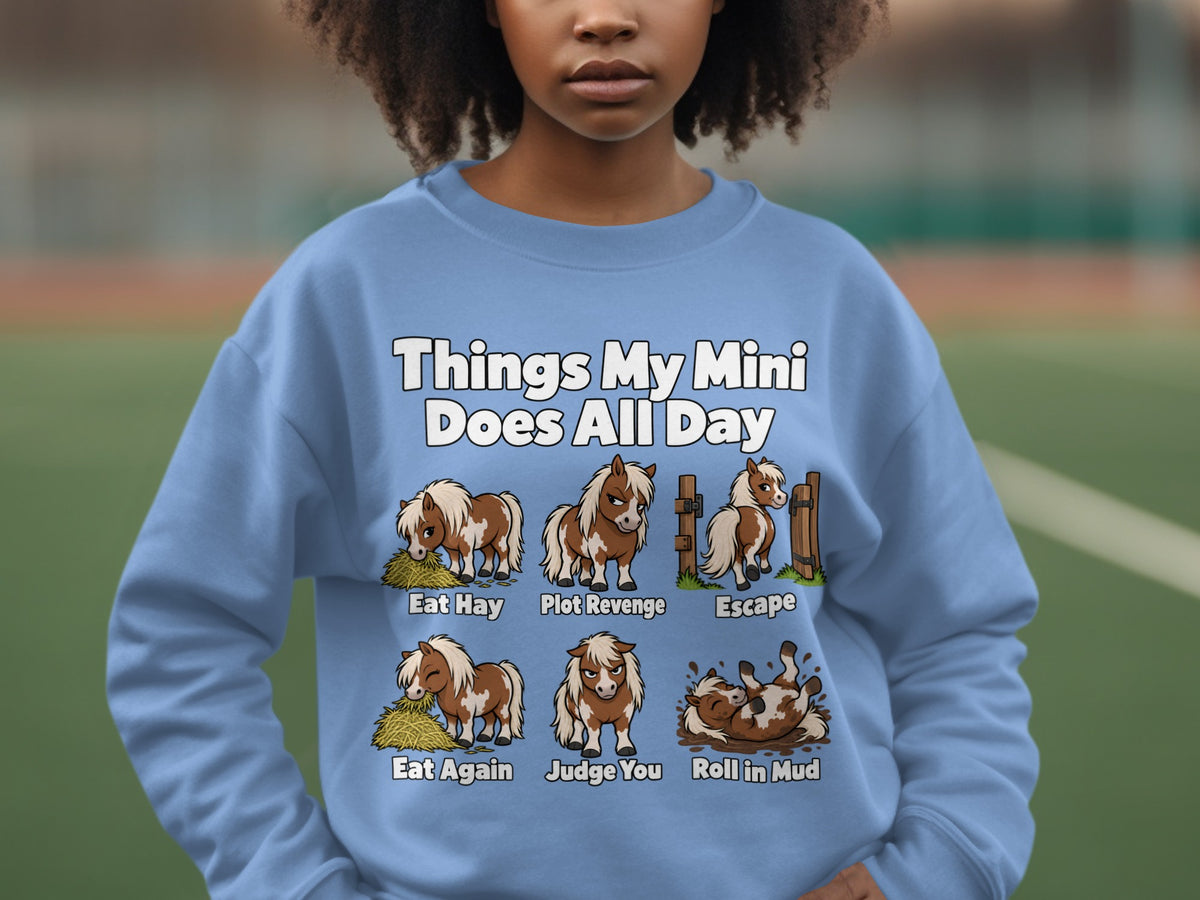 A person wearing a light blue sweatshirt printed with cartoon illustrations of miniature horses and humorous captions.