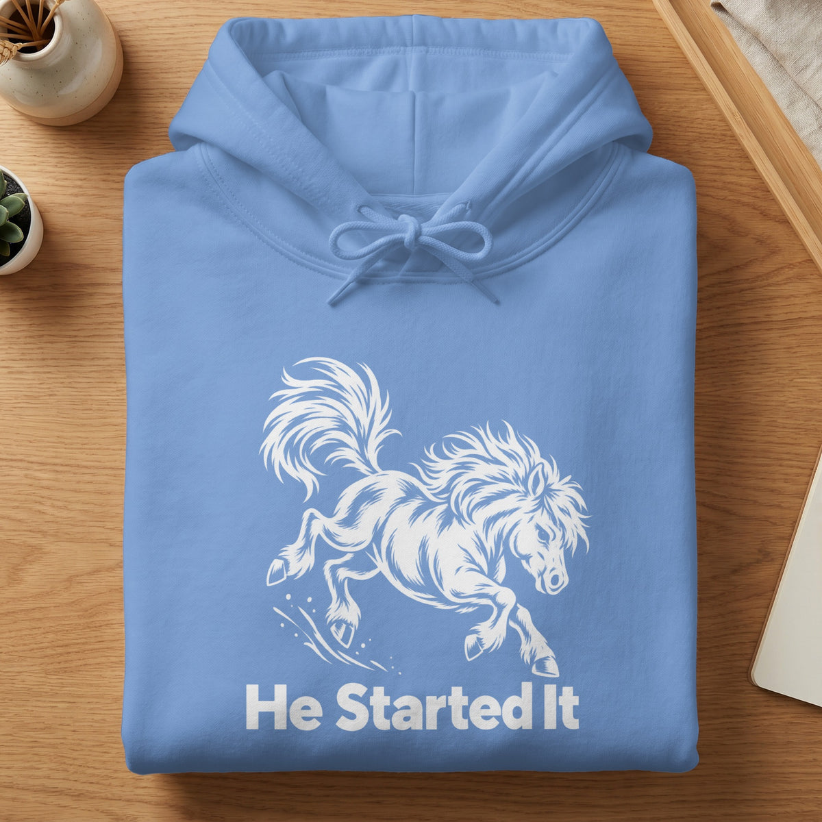 A folded light blue hoodie featuring a white graphic of a running horse and the text He Started It.
