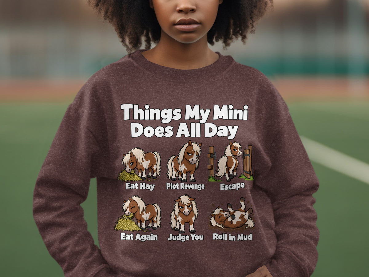 A person wearing a brown crewneck sweatshirt featuring illustrations and humorous text about miniature horses.