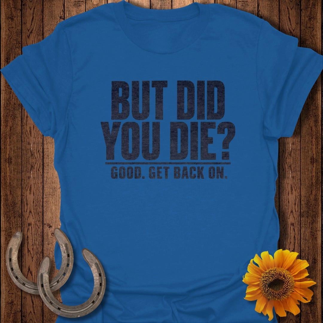 A blue t-shirt printed with the text "BUT DID YOU DIE? GOOD. GET BACK ON." lies on a wooden background next to two horseshoes and a yellow sunflower.