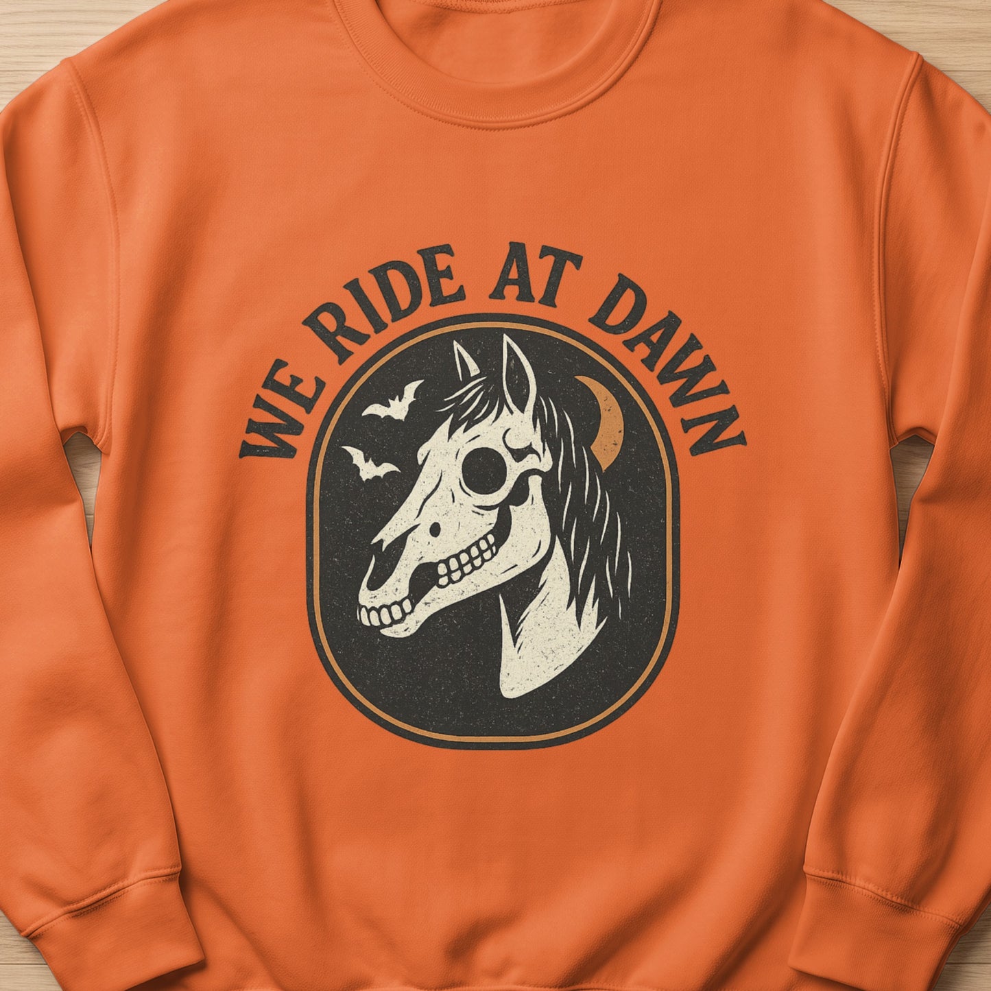 Orange sweatshirt with black circle graphic featuring horse skull, bats, moon, and text "WE RIDE AT DAWN."