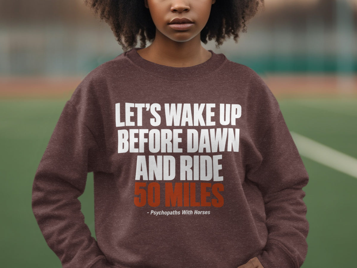A person wearing a brown sweatshirt with white and orange text that reads Let's Wake Up Before Dawn And Ride 50 Miles.