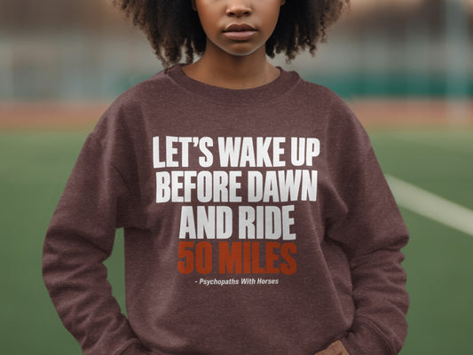 A person wearing a brown sweatshirt with white and orange text that reads Let's Wake Up Before Dawn And Ride 50 Miles.