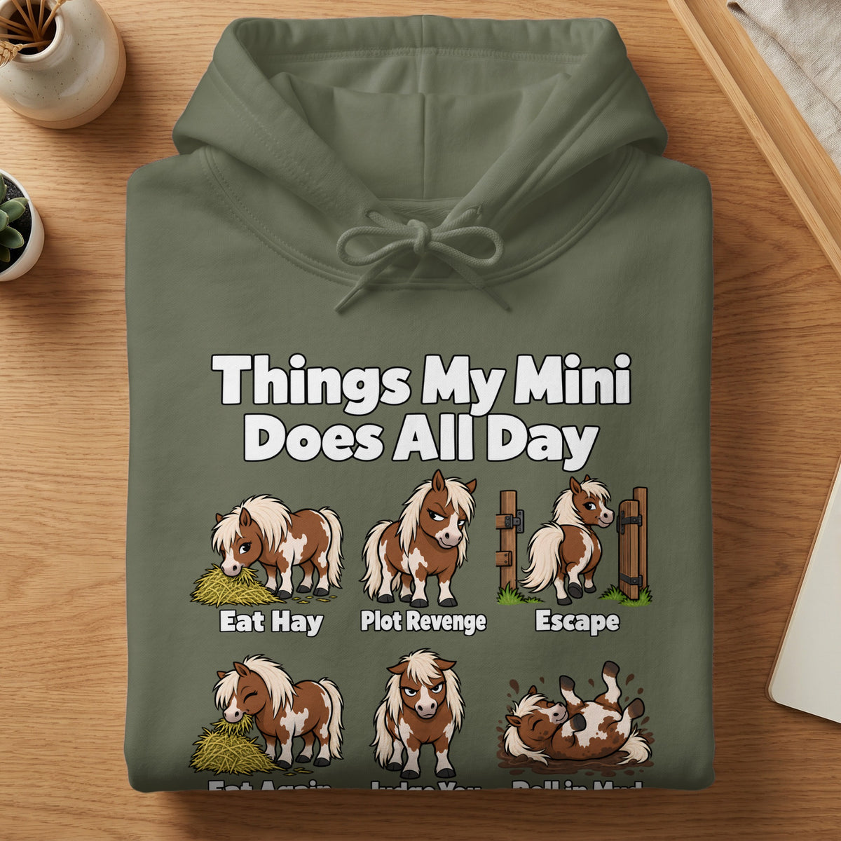 Olive green hooded sweatshirt folded flat on a wooden surface, featuring cartoon illustrations and text about mini horses.