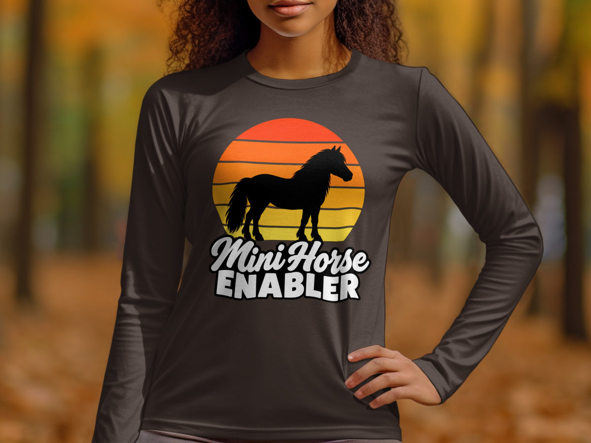 A person wearing a long-sleeve charcoal gray shirt featuring a horse silhouette over a sunset and the text Mini Horse Enabler.