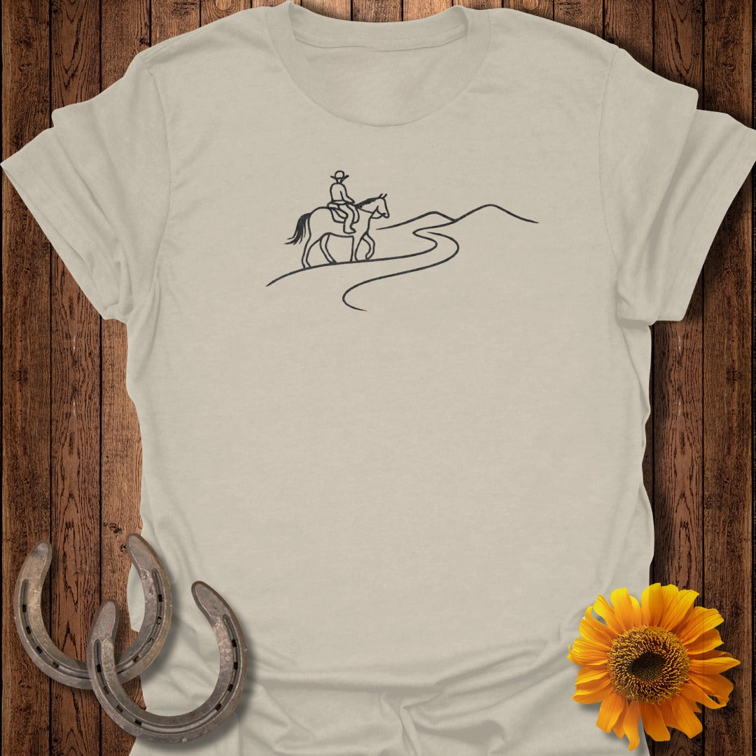 Light tan t-shirt with a black line drawing of a cowboy riding a horse on a winding path.