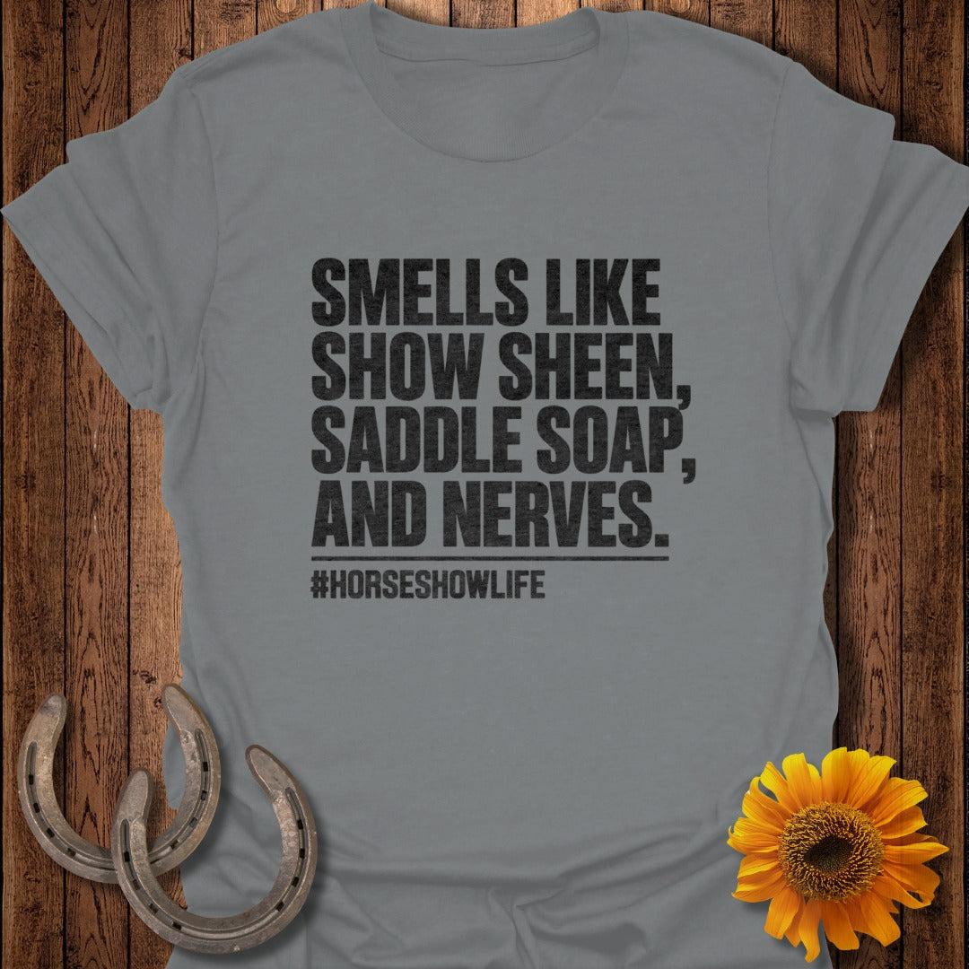 A gray t-shirt with black text reading "Smells like show sheen, saddle soap, and nerves. #horseshowlife" laid on wood with horseshoes and a sunflower.