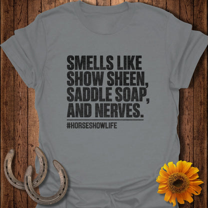 A gray t-shirt with black text reading "Smells like show sheen, saddle soap, and nerves. #horseshowlife" laid on wood with horseshoes and a sunflower.