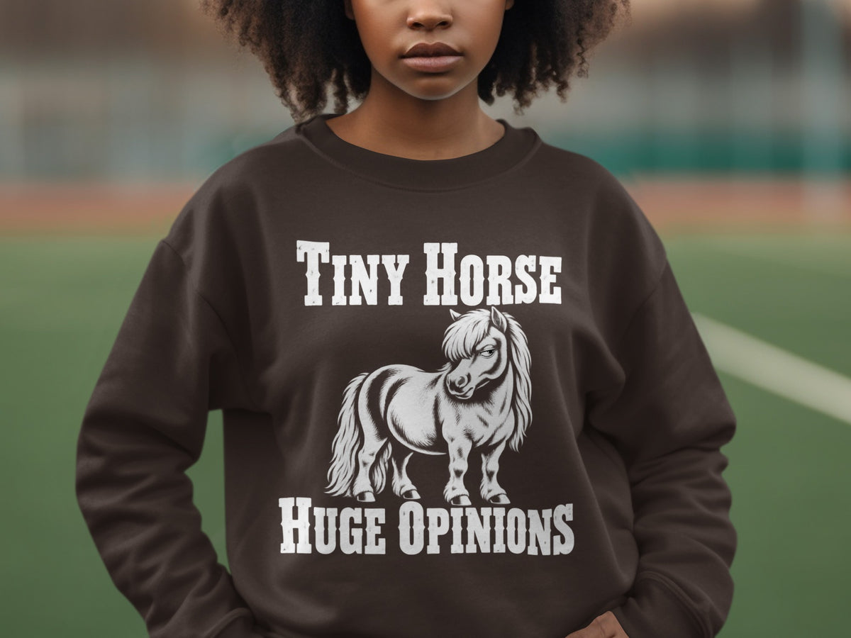 A person wearing a brown crewneck sweatshirt featuring a white graphic of a pony and the text Tiny Horse Huge Opinions.