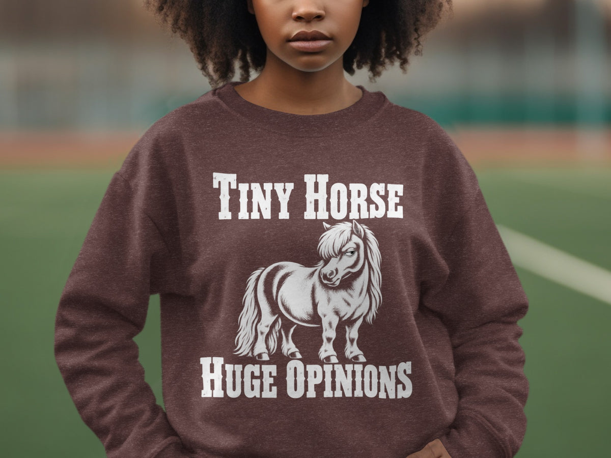 A person wearing a brown crewneck sweatshirt featuring a graphic of a pony and the text Tiny Horse Huge Opinions.