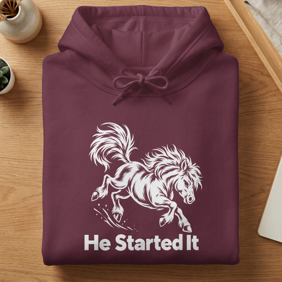 A folded maroon hoodie with a white graphic of a bucking horse and the text He Started It.