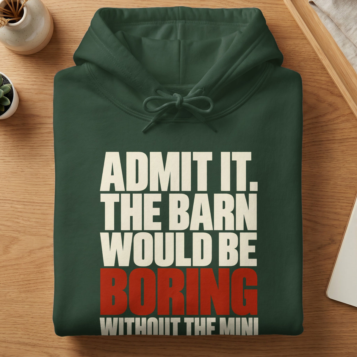 A folded dark green hoodie on a wooden surface with white and red text reading ADMIT IT. THE BARN WOULD BE BORING WITHOUT THE MINI.