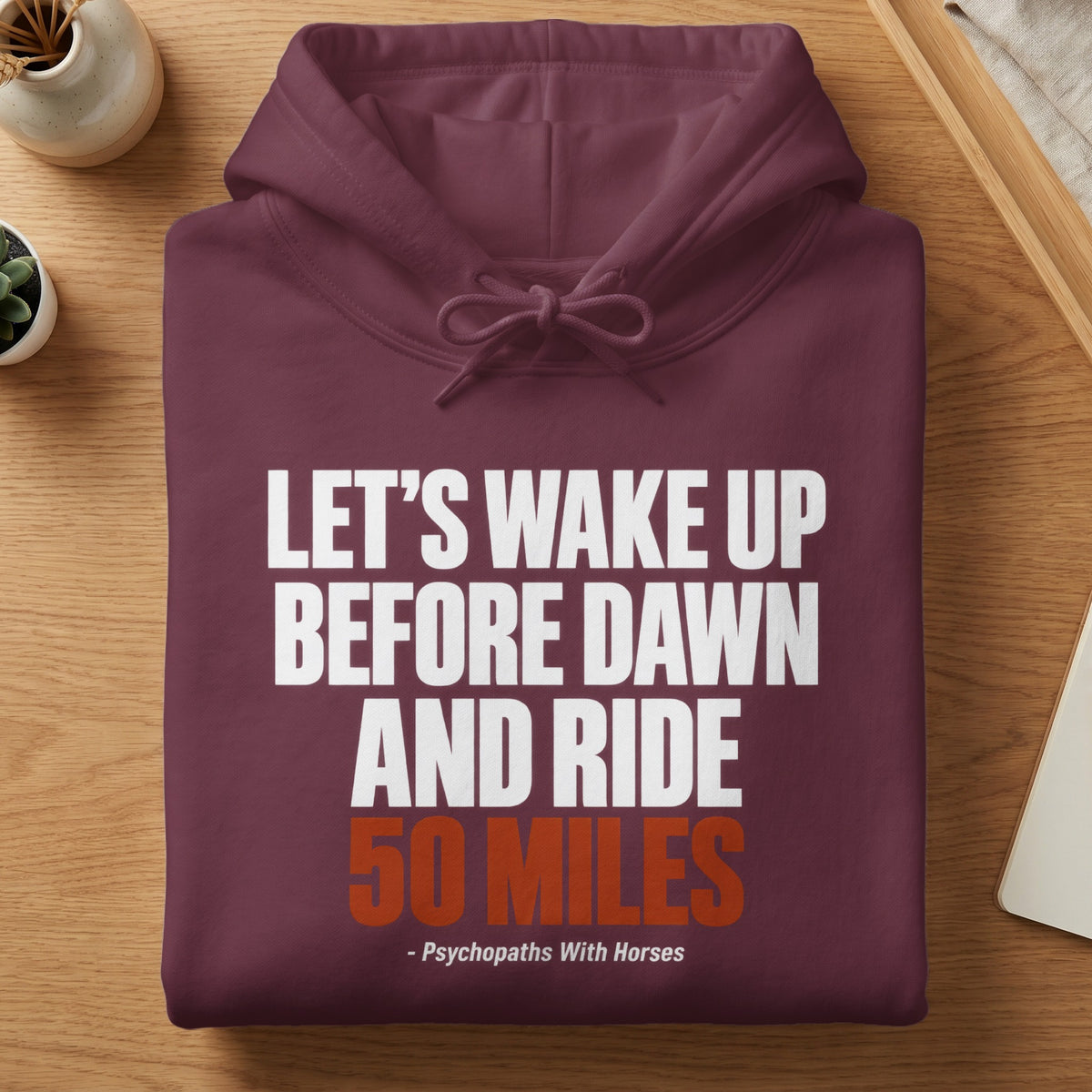 Folded maroon hoodie on a wooden table, featuring white and orange text that reads "Let's wake up before dawn and ride 50 miles - Psychopaths With Horses".
