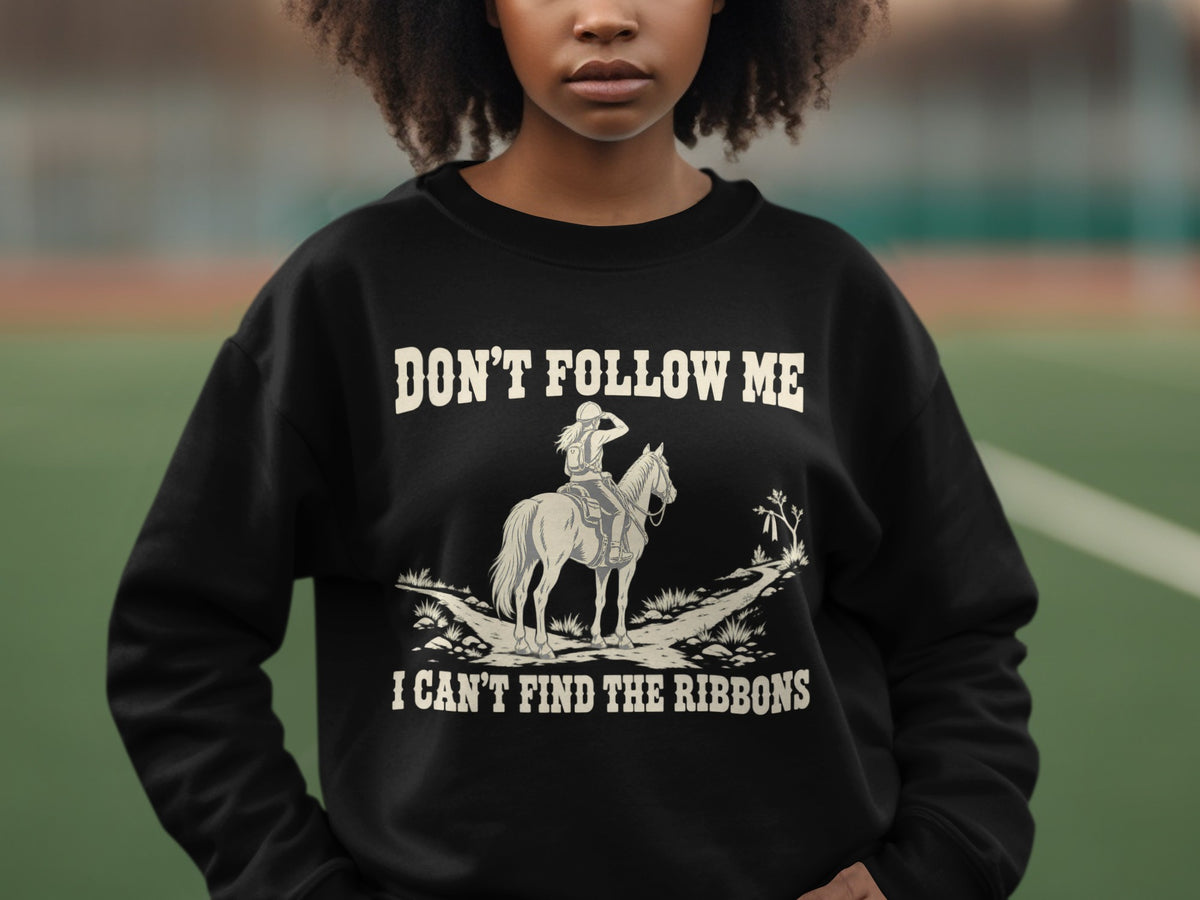 A person wearing a black sweatshirt printed with the text DON'T FOLLOW ME I CAN'T FIND THE RIBBONS and an illustration of an equestrian on a horse.