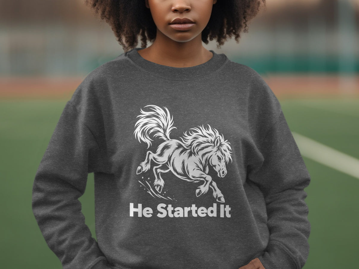 A person wearing a dark grey crewneck sweatshirt featuring a white graphic of a running horse and the text He Started It.