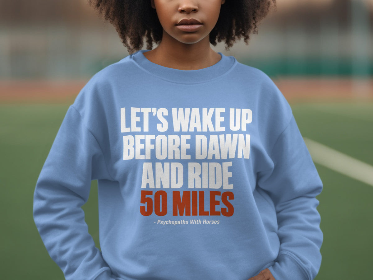 A person wearing a light blue crewneck sweatshirt with the text "LET'S WAKE UP BEFORE DAWN AND RIDE 50 MILES" printed on the chest.