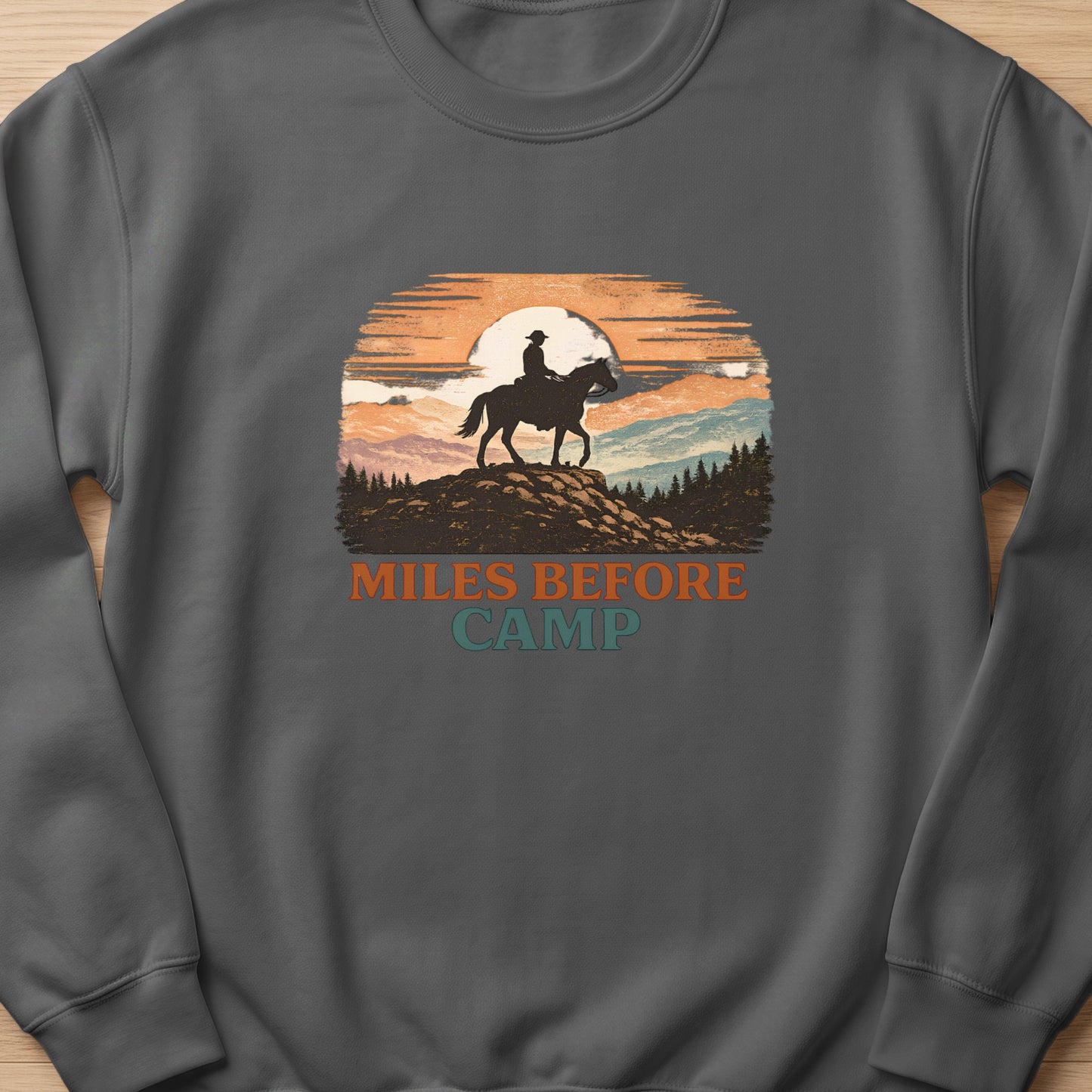 Sweatshirt with a graphic of a cowboy on horseback at sunset with text "Miles Before Camp."