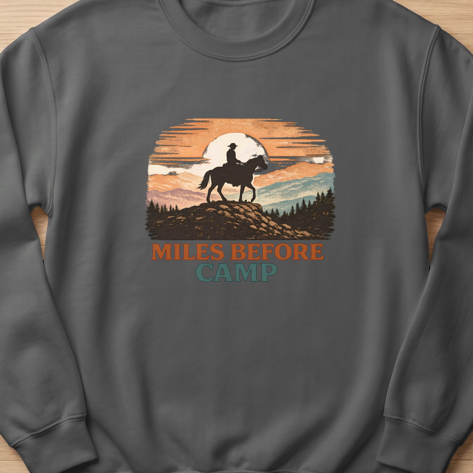 Sweatshirt with a graphic of a cowboy on horseback at sunset with text "Miles Before Camp."
