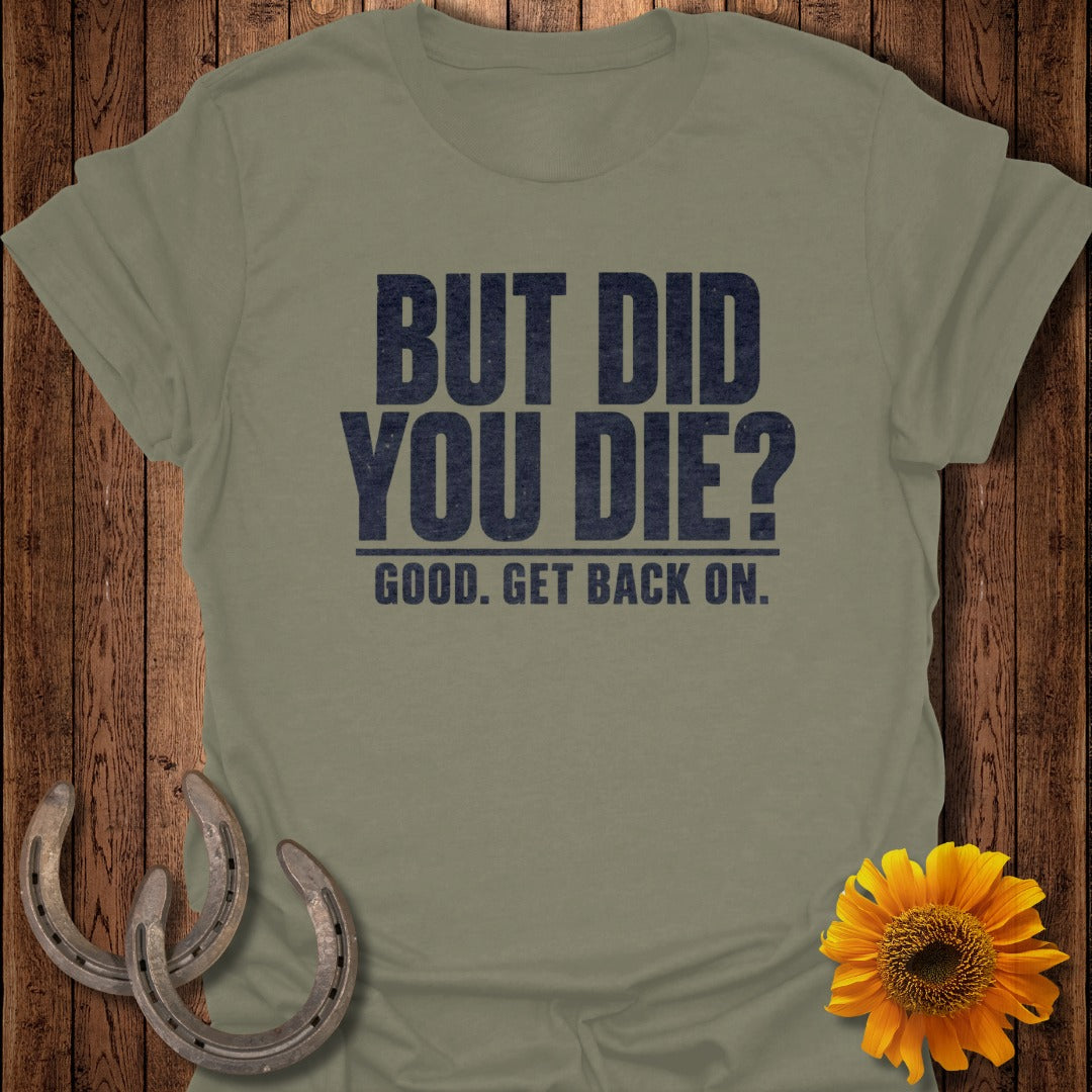 An olive green t-shirt with black text reading "BUT DID YOU DIE? GOOD. GET BACK ON." on a wooden background with horseshoes and a sunflower.