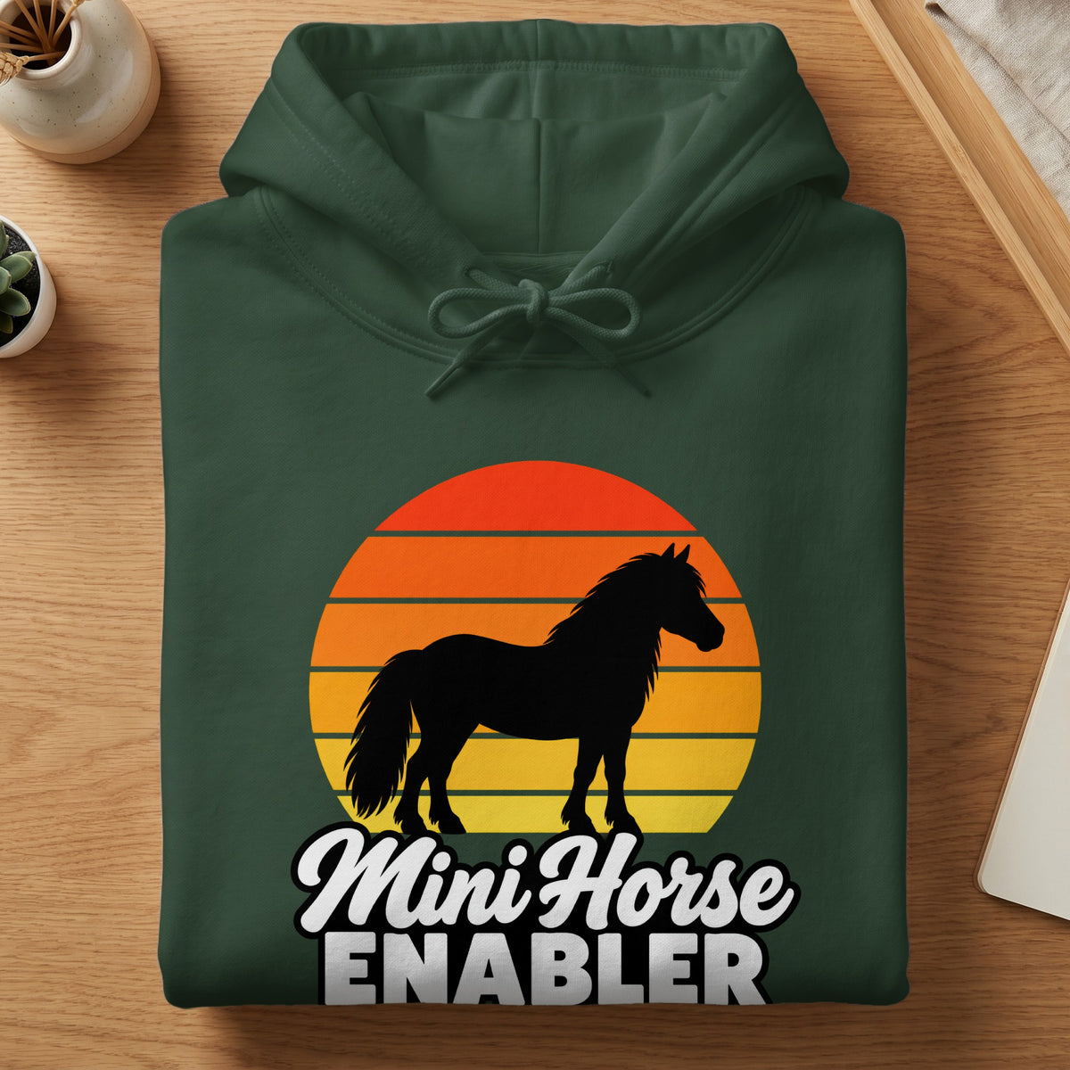 A folded dark green hoodie featuring a horse silhouette over a striped sunset and the text Mini Horse ENABLER.