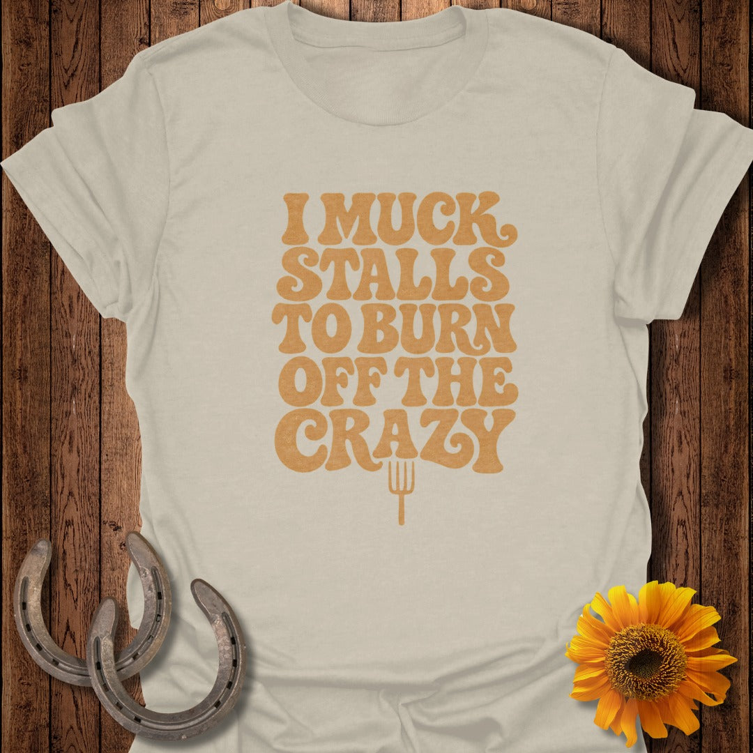A beige crewneck t-shirt featuring the orange text I MUCK STALLS TO BURN OFF THE CRAZY.