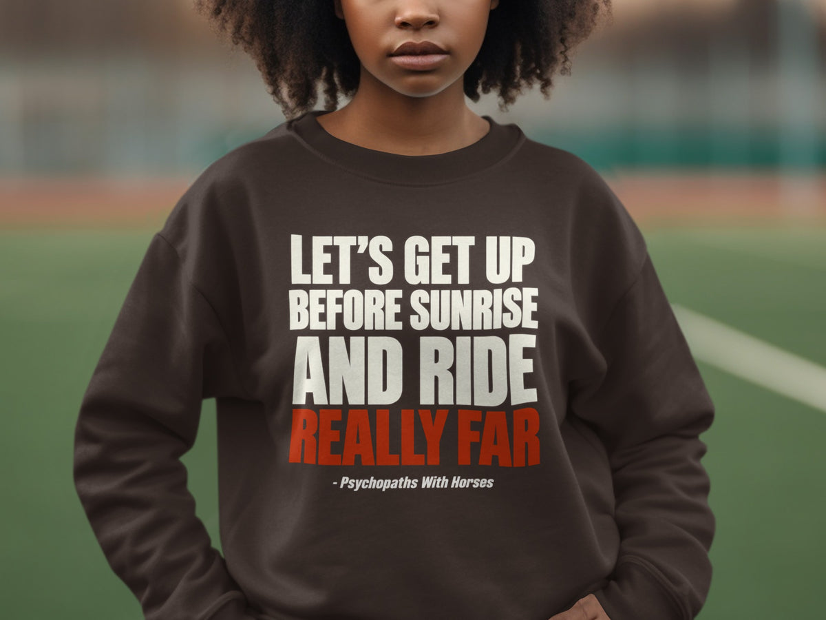 A person wears a brown crewneck sweatshirt featuring white and red text stating, "Let's Get Up Before Sunrise And Ride Really Far - Psychopaths With Horses."