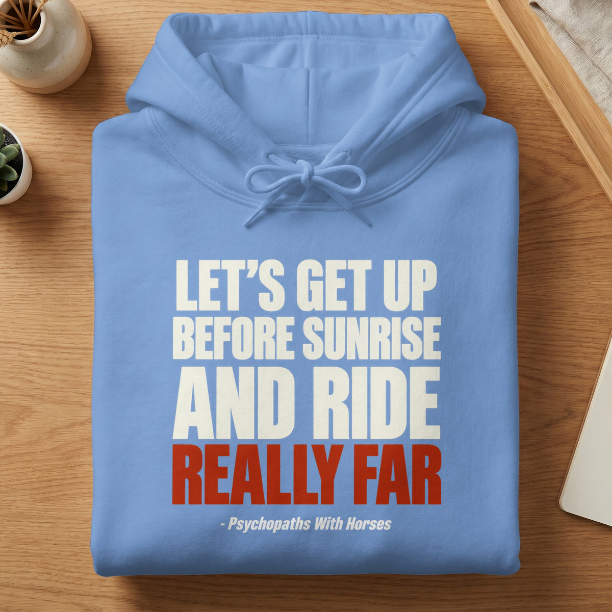 A folded light blue hoodie featuring white and red text stating, Let's get up before sunrise and ride really far.