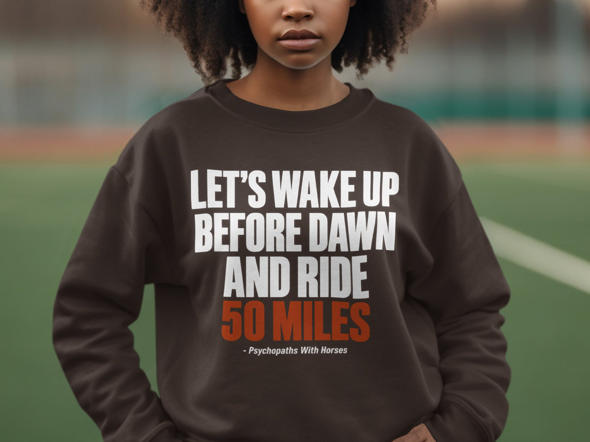 A person wearing a brown crewneck sweatshirt featuring white and orange text that reads Let us wake up before dawn and ride 50 miles.