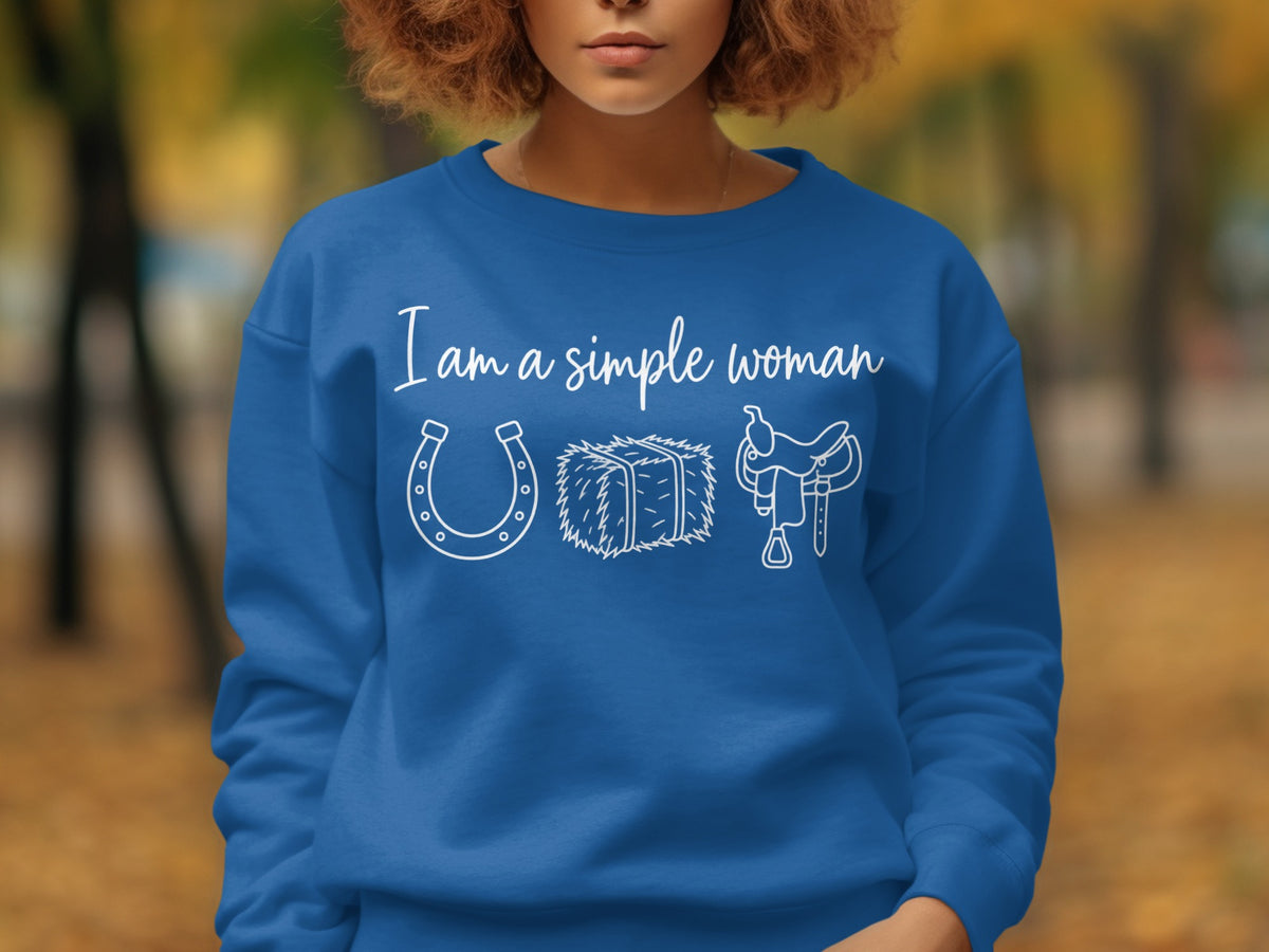 Blue sweatshirt with white text "I am a simple woman" above horseshoe, hay bale, and saddle icons.